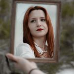 A young redhead woman holding a mirror outdoors, showcasing her reflection with red lips.